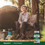 A middle-aged woman laughs while sitting in a camping chair beside a tent, holding a bottle of African Mango & Green Tea.