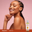 A young woman smiles with closed eyes and hand on chin next to an Arnica Massage Oil bottle.