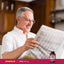 An older man smiling as he reads a newspaper next to a bottle of DMAE.
