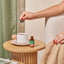 A woman's hand uses a dropper to add Lion's Mane Liquid Drops to coffee on a wooden table.