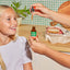 A young blonde girl smiles at an adult offering Fungies Mushroom Liquid Drops beside a lunchbox with a sandwich.