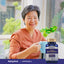 Elderly Asian woman smiling, holding a mug while sitting next to a bottle of Huperzine A.