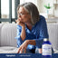 An older woman sits on a couch, holding a pen and looking down, with a bottle of L-Methylfolate beside her.