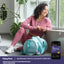 A woman in pink athletic wear sits on a yoga mat by a window, smiling, with a Magnesium Drink Mix.