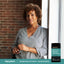 A middle-aged woman smiles by a window with an Ashwagandha + Mag Glycina supplement bottle beside her.