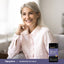 A middle-aged woman smiles while relaxing on a couch with a jar of Magnesium Glycinate powder beside her.