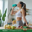 Woman drinking from a green shaker bottle in a kitchen, with an Oil of Oregano bottle visible on the counter.