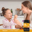 Child smiling while being fed with a spoon by an adult at a kitchen table, with a Piping Rock Kids Multi + Probiotic bottle nearby