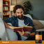 Lifestyle image of man relaxing on a couch while reading a book, with Piping Rock Sublingual B12 Methylcobalamin supplement bottle nearby
