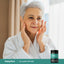 Adult touching facial skin while wearing a white robe with a Piping Rock Ultra Collagen Peptides Complex bottle nearby.