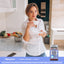 A young woman smiles while holding a teacup in a kitchen next to a bottle of Stress Support Gummies.