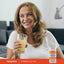 Adult smiling while holding a glass of orange juice, with a Piping Rock Absorbable Vitamin C Plus Zinc, 180 softgels bottle shown in the bottom corner.