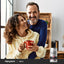 tTwo adults smiling and holding a red mug together indoors with a Piping Rock Red Yellow Black Maca supplement bottle displayed in the bottom right corner