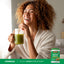 Young woman laughing and drinking a green smoothie from a jar, with a jar of Organic Greens & Fruits Plus beside her.