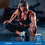 Adult stretching while seated on a gym floor, with a Lindberg Varsity Pack 2 Health, Fitness & Athletics supplement container nearby