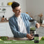 A young man blends a green smoothie in the kitchen with a Spirulina bottle on the counter.