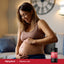 Pregnant adult smiling while holding their baby bump indoors, with a Piping Rock Prenatal DHA bottle nearby