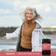 Adult woman smiling outdoors near the water wearing a white jacket and brown sweater, with a bottle of Piping Rock Primal Beef Organs For Women displayed in the lower right corner.