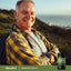 An older man with a backpack smiles on a hillside, holding a bottle of Sea Buckthorn Oil.