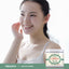 A young adult woman smiles and touches her cheek next to a jar of Lanolin Cream.