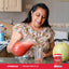 A woman pours a red drink into a glass, with an Organic Reds Plus bottle and a watermelon next to her.