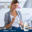 Young woman enjoys yogurt in a kitchen, seated next to a bottle of Serrapeptase, portraying a healthy lifestyle.