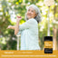 A senior woman stretches her arm while exercising outdoors, standing beside a Joint Support bottle.