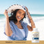 A young woman smiles and holds a straw hat on a beach, next to a Pancreatin Enzyme bottle.