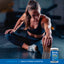 A young adult woman stretches next to a Varsity Pack 2 jar, promoting a healthy lifestyle.