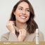 Young adult woman smiling while holding her sweater collar, next to a Vitamin E Moisturizing Stick on display.