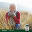 Older woman in winter clothing crouches in tall grass, looking at the camera, beside a Yucca Root bottle.